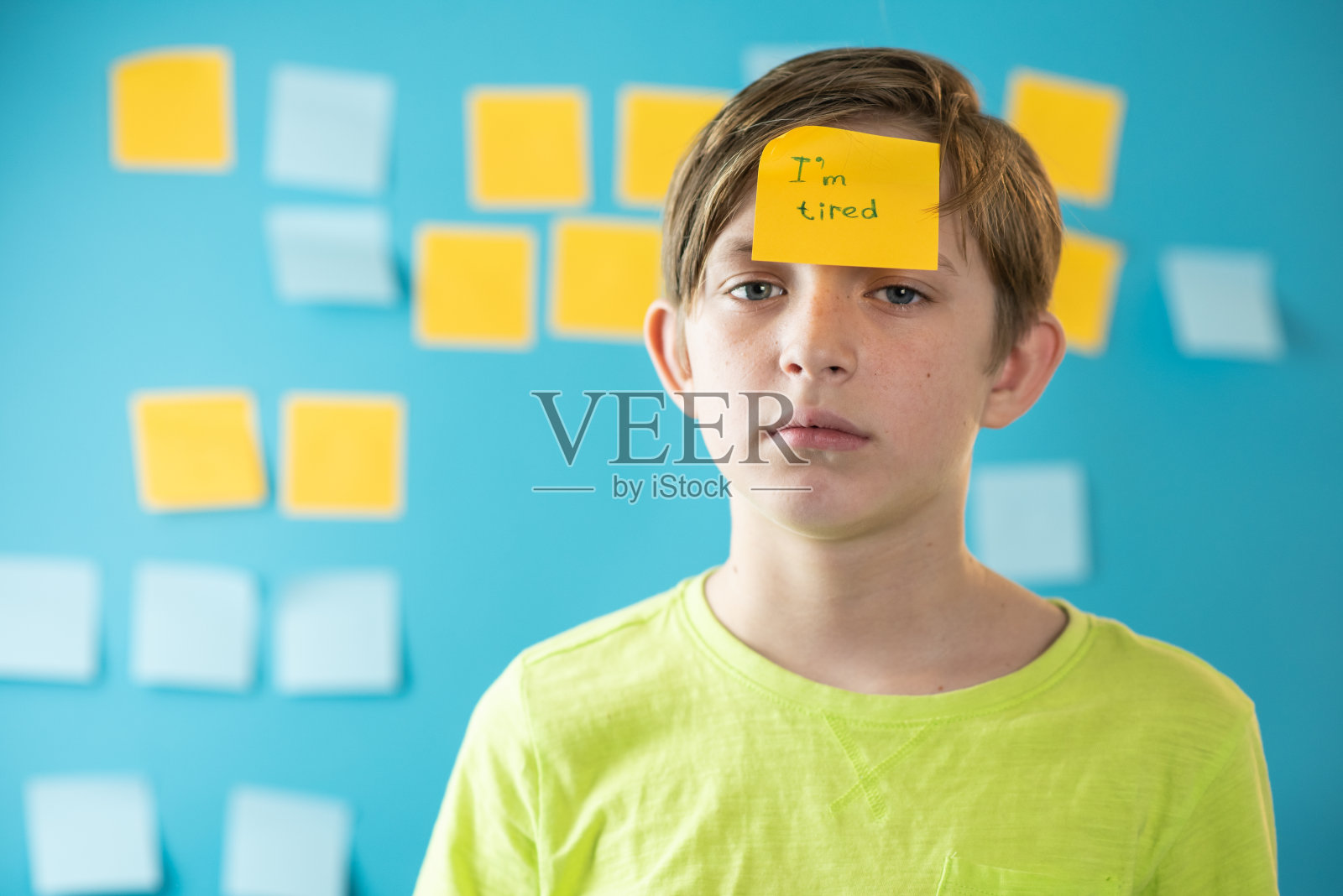 一个疲惫的11岁男孩站在贴有贴纸的蓝色墙壁前。额头上贴着“我累了”贴纸的严肃儿童照片摄影图片