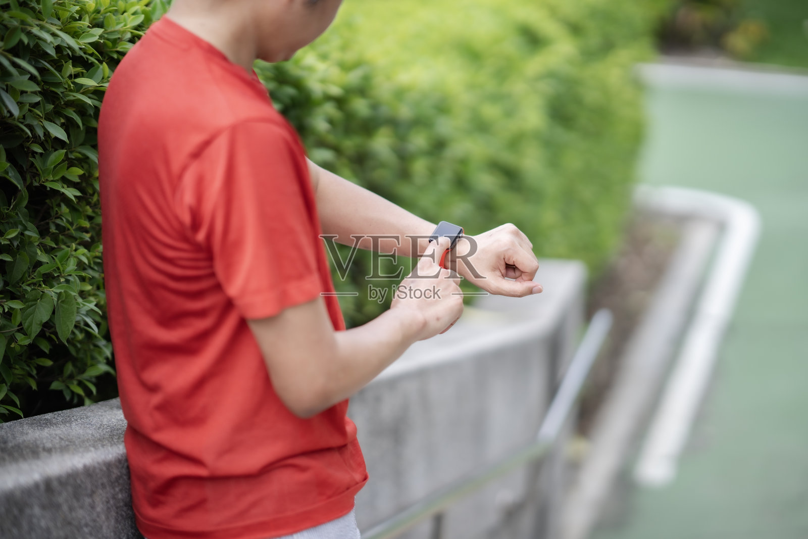 关注智能手表,一位中年男子站在公园的后面,触摸并看着红色的智能手表。照片摄影图片