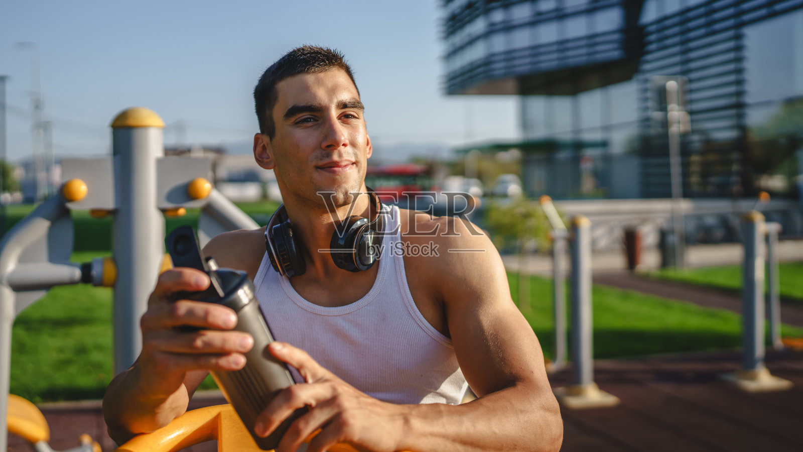 一名白人男子年轻男运动员在公园户外健身房户外训练时刹车,手里拿着补品摇瓶快乐自信强的复制空间照片摄影图片