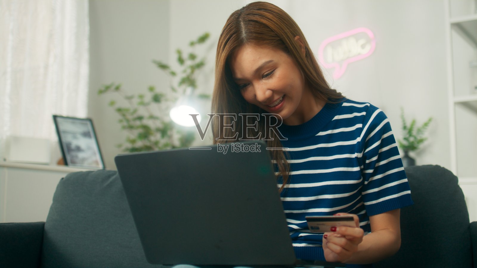 年轻的亚洲女子手持信用卡,用笔记本电脑进行网上购物,女子在家里的客厅里享受着购买衣服的乐趣照片摄影图片