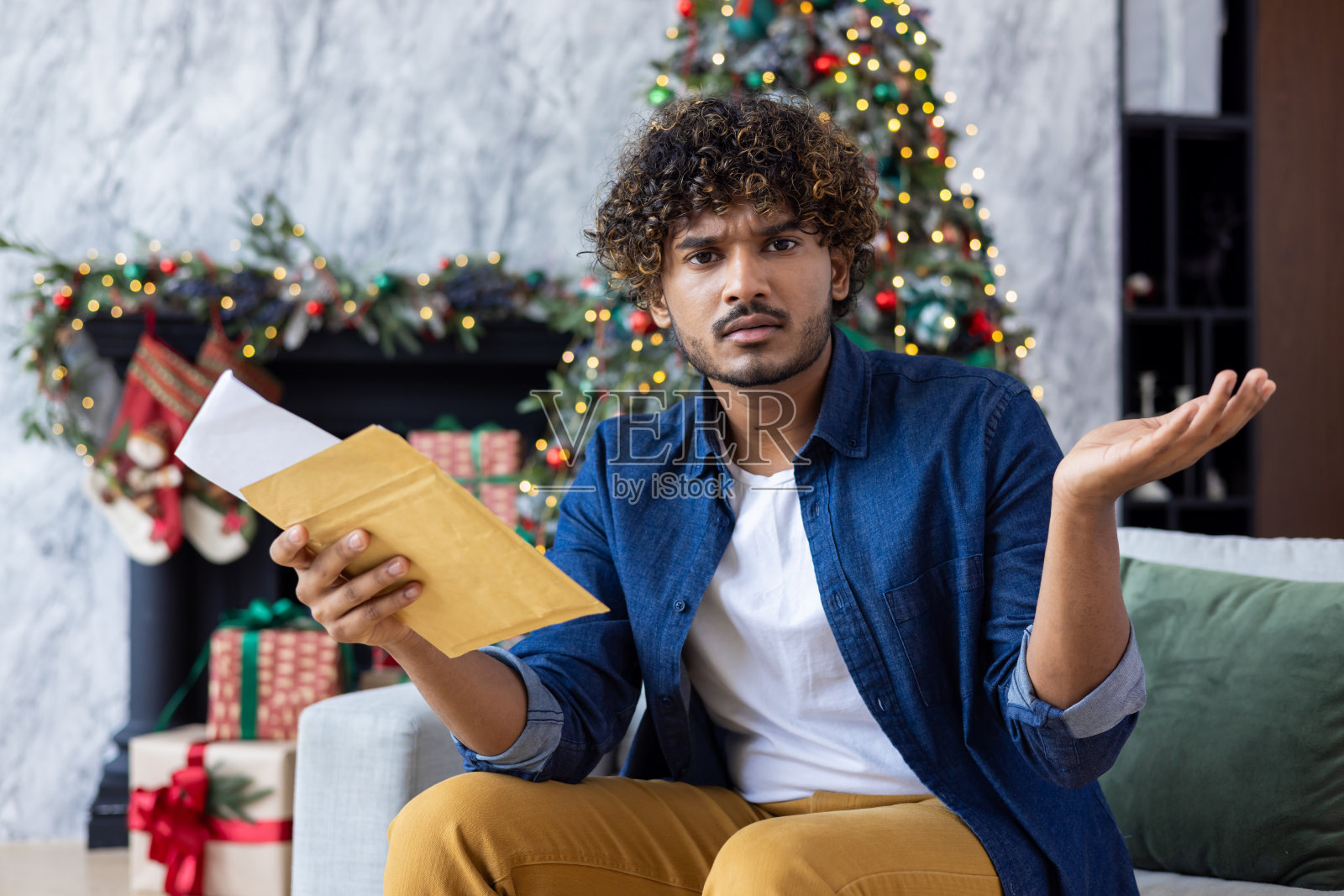 一个失望和沮丧的年轻印度男子的肖像,他在新年假期收到了坏消息。坐在家里的沙发上,看着相机,摊开双手照片摄影图片