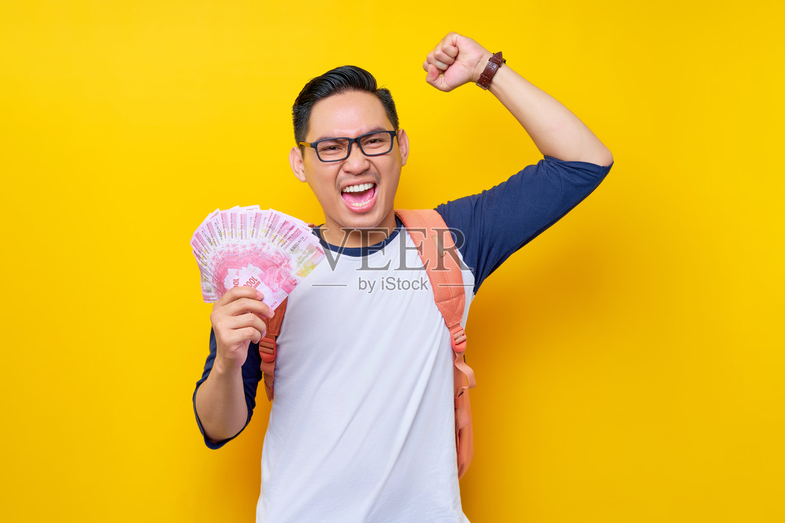 兴奋的年轻亚洲学生男子穿着休闲服,戴着眼镜,背包里拿着现金,庆祝获胜者被孤立在黄色背景中。高中教育大学学院理念照片摄影图片