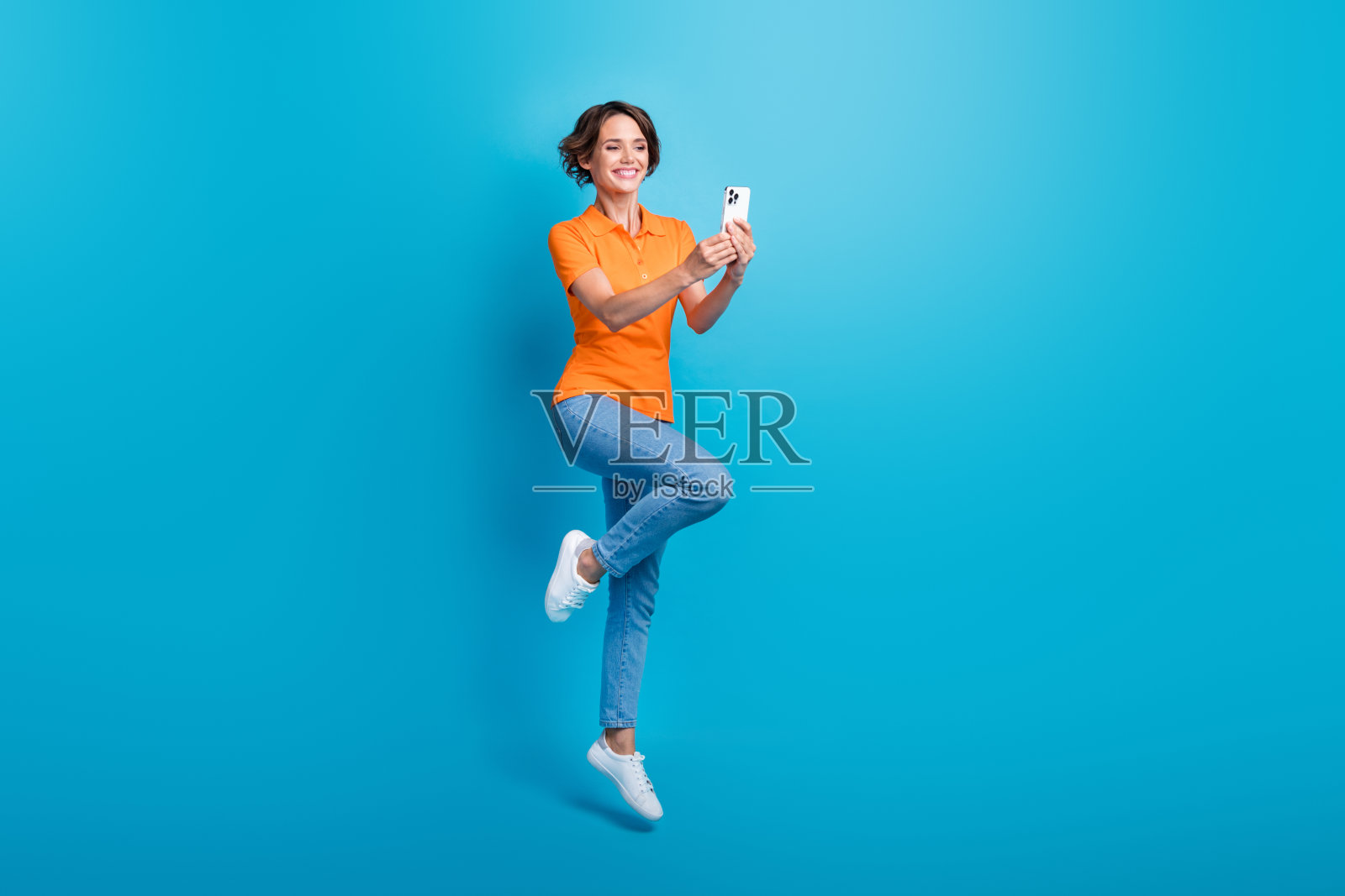 可爱乐观的女人全身照片,穿着橙色t恤,时尚牛仔裤,在蓝色背景下跳着自拍照片摄影图片
