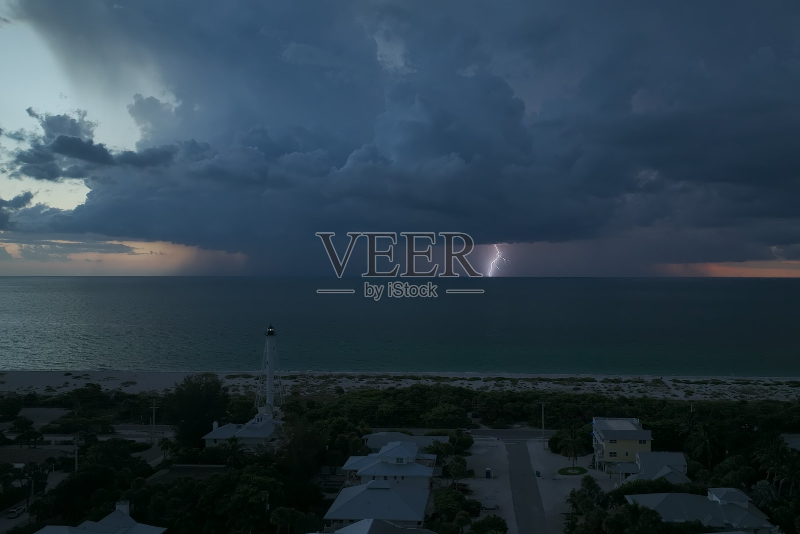 海边的小镇,黑暗的房屋和矗立在海边的高大灯塔,在暴风雨的夜晚闪烁着灯光,为船只导航。海水上有闪电的雷暴,对船只构成危险照片摄影图片