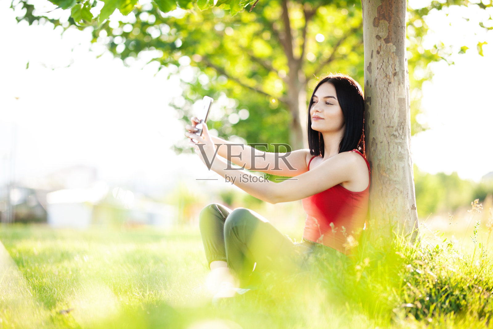 快乐的深棕色头发的女孩在树下拿着她的智能手机照片摄影图片