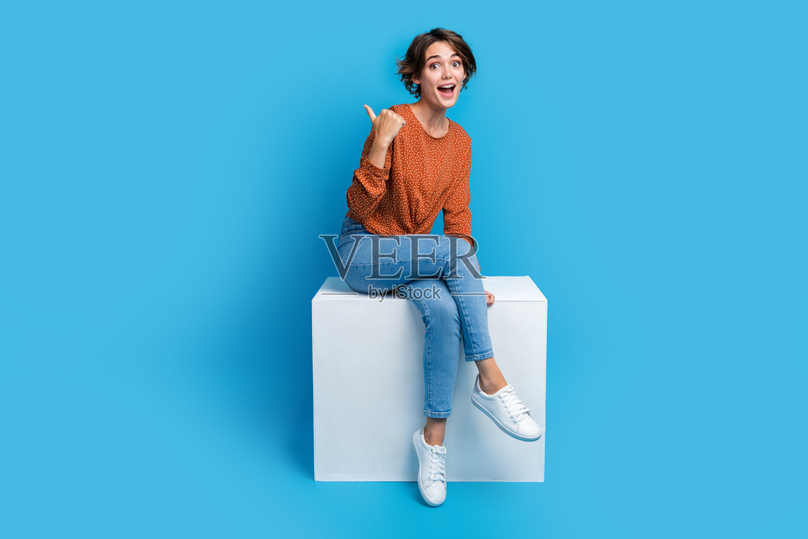 漂亮的年轻女孩的全身照片点空印象深刻,坐在讲台上穿时髦的棕色衣服孤立在蓝色的背景照片摄影图片