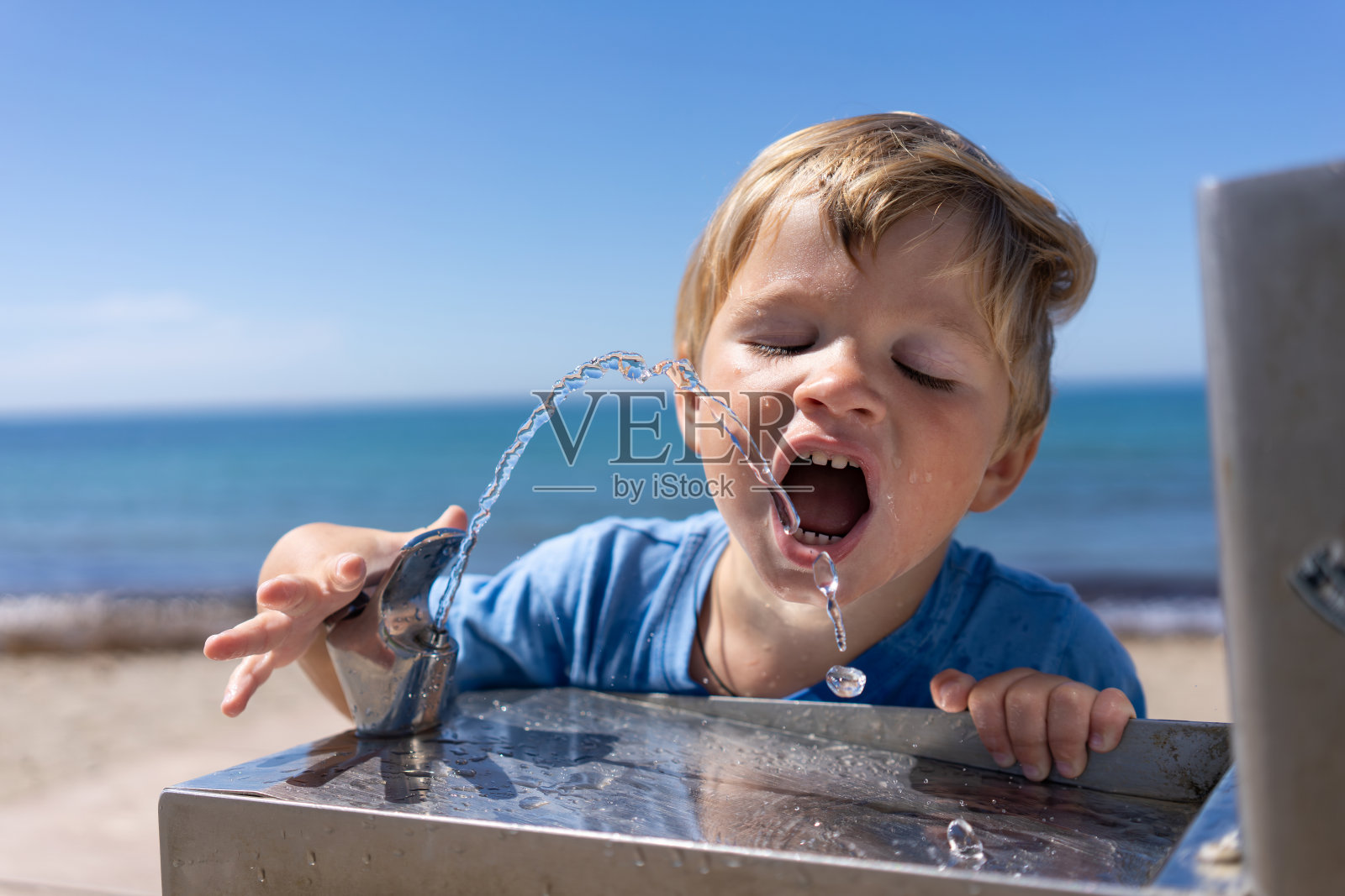 3岁的金发男孩在夏天从海滩上的饮水机里喝水照片摄影图片
