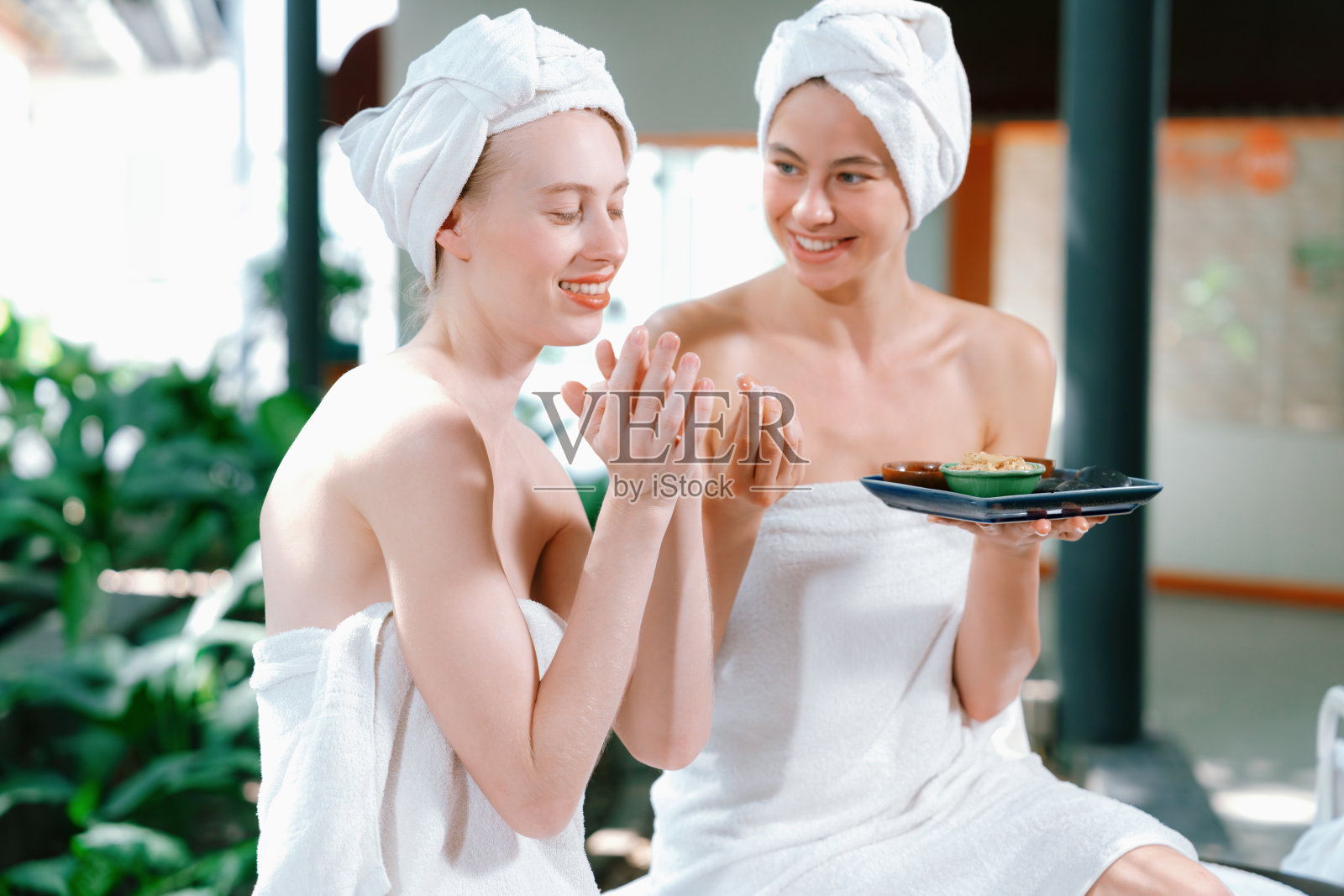 两个漂亮的女人裹着毛巾,一边拿着草药碗一边咯咯地笑。宁静。照片摄影图片