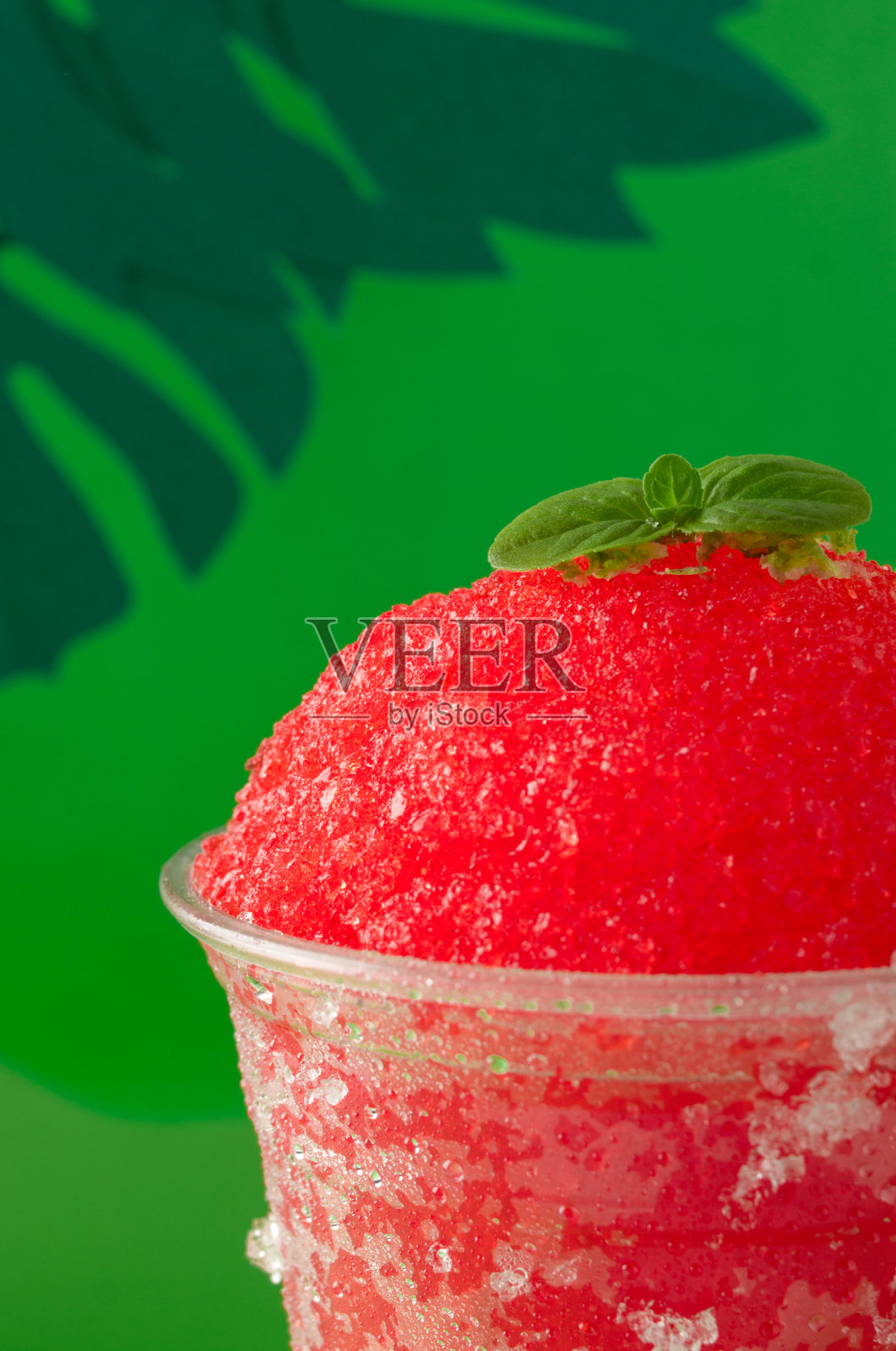 草莓和樱桃红色水果味的刨冰或柠檬冰沙,装在一个透明的一次性杯子里,用绿色的纸做背景,用绿色的纸叶装饰。照片摄影图片