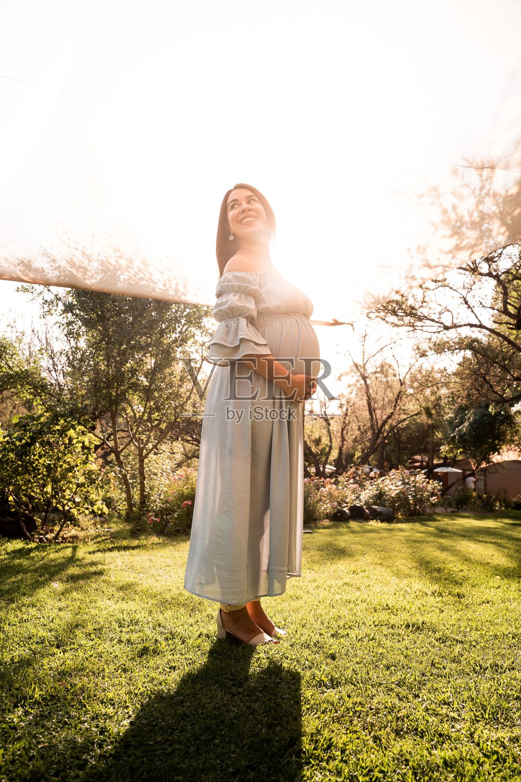 一名穿着裙子的拉丁孕妇站在郁郁葱葱、阳光明媚的公园里,双手轻轻地抚摸着她的肚子照片摄影图片