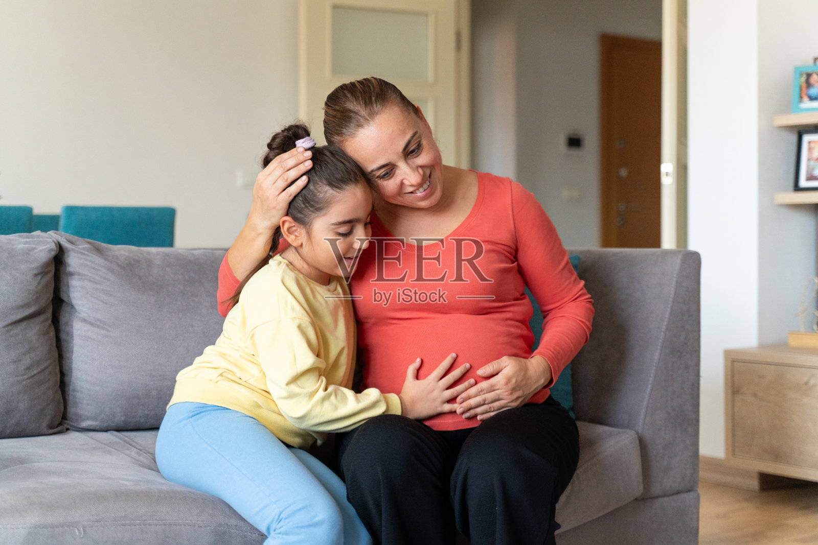 小女孩和她怀孕的妈妈坐在家里,深情地拥抱在一起照片摄影图片