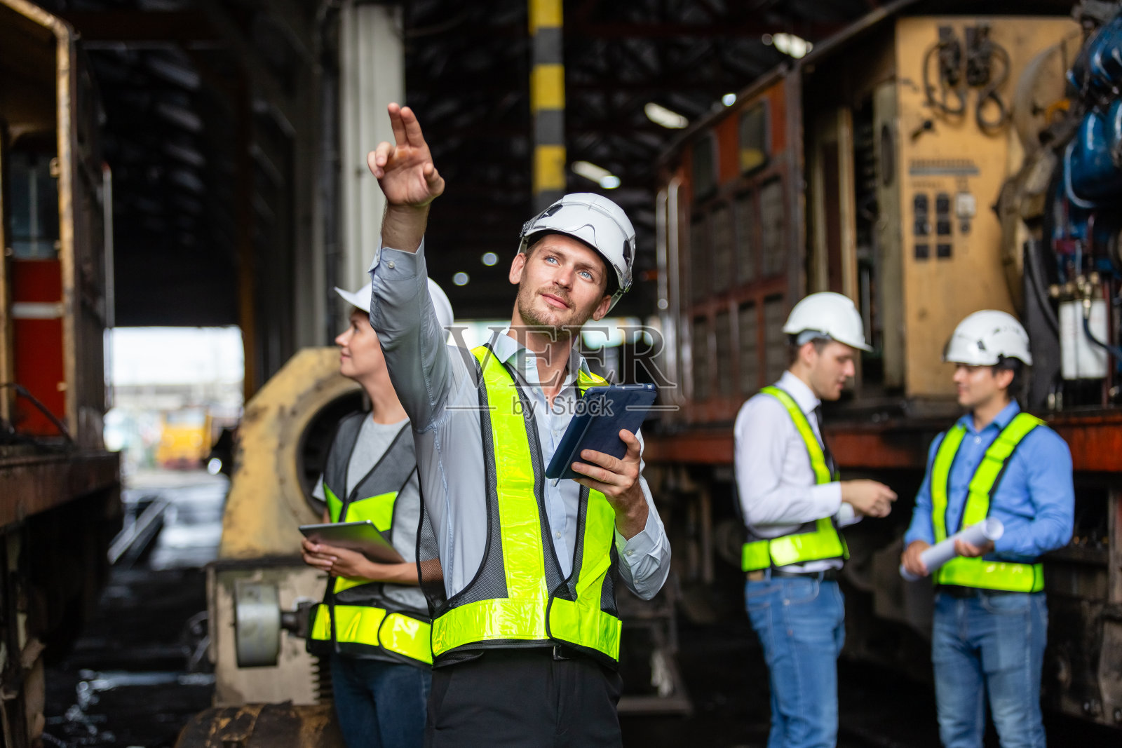 一名铁路技术工程师,身穿安全制服,头戴安全帽,手持平板,站在现场火车站,具有团队合作背景。照片摄影图片