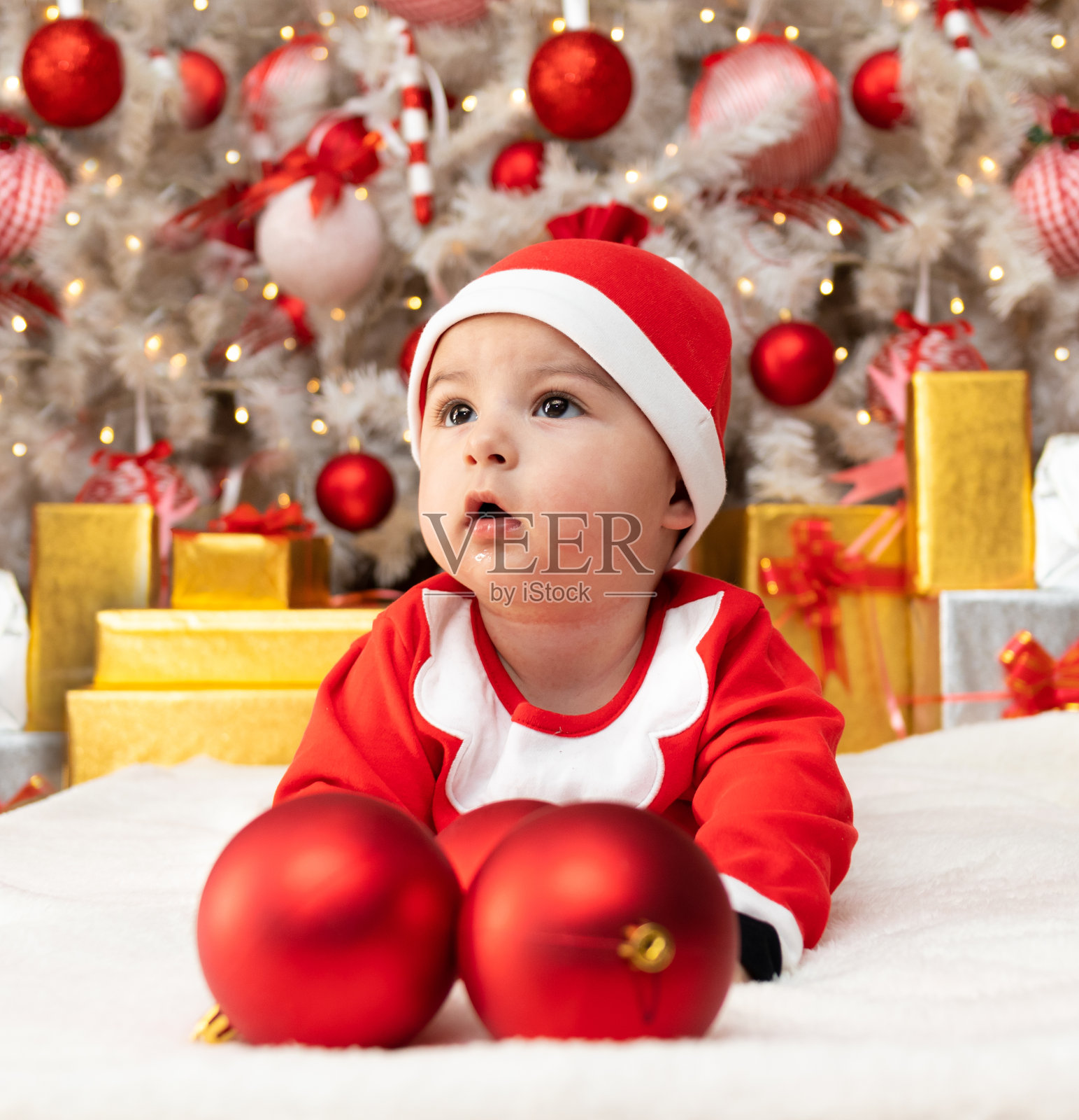 可爱的宝宝戴着红色的圣诞帽,穿着圣诞服,在地板上玩着圣诞灯和礼物盒照片摄影图片