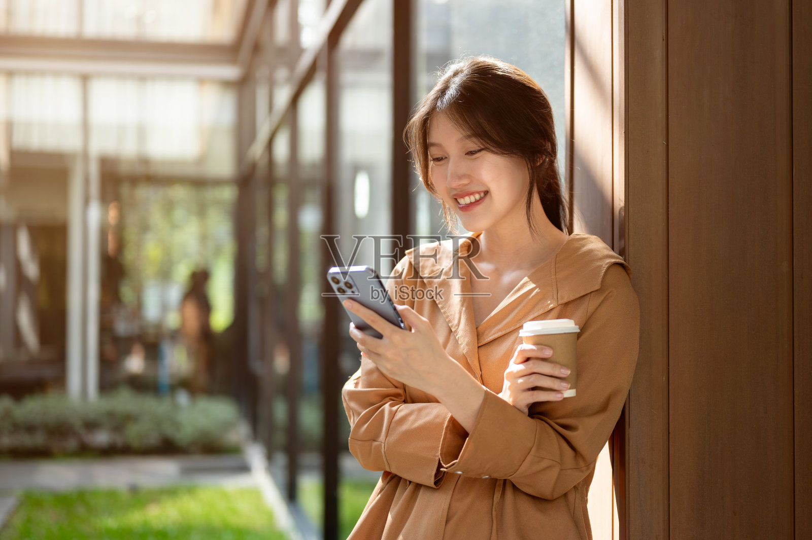 一位迷人的亚洲女性倚在户外的墙上,一边用智能手机一边喝着咖啡。照片摄影图片