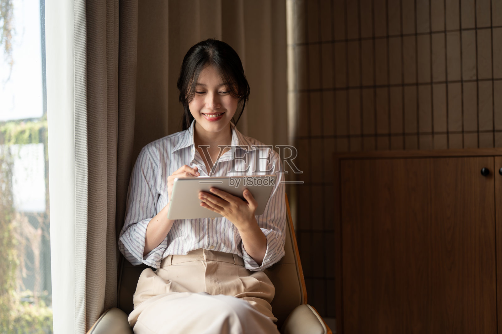 一位迷人的亚洲女性坐在室内靠窗的地方玩着她的平板电脑。照片摄影图片