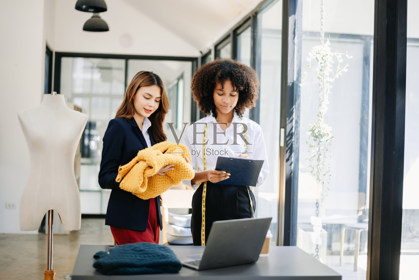 不同的女性时装设计师们脖子上戴着“裁缝厘米”,手里拿着平板电脑和智能手机。照片摄影图片