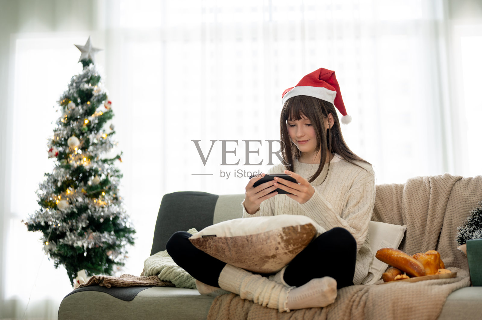 圣诞节当天,一位戴着圣诞老人帽子的快乐女子躺在沙发上享受着手机游戏。照片摄影图片