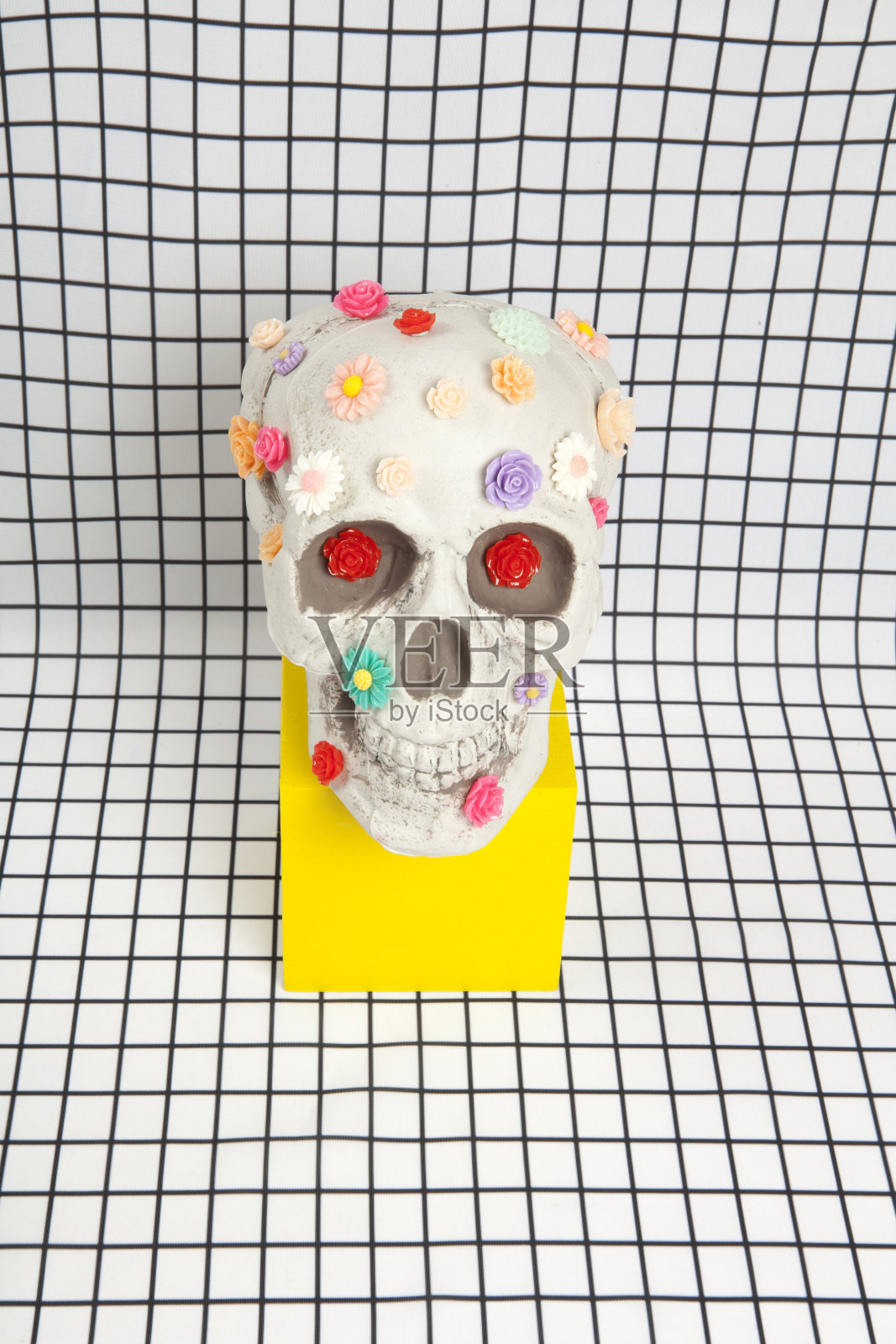 在白色方形瓷砖图案上,一个头骨上覆盖着多朵塑料花照片摄影图片
