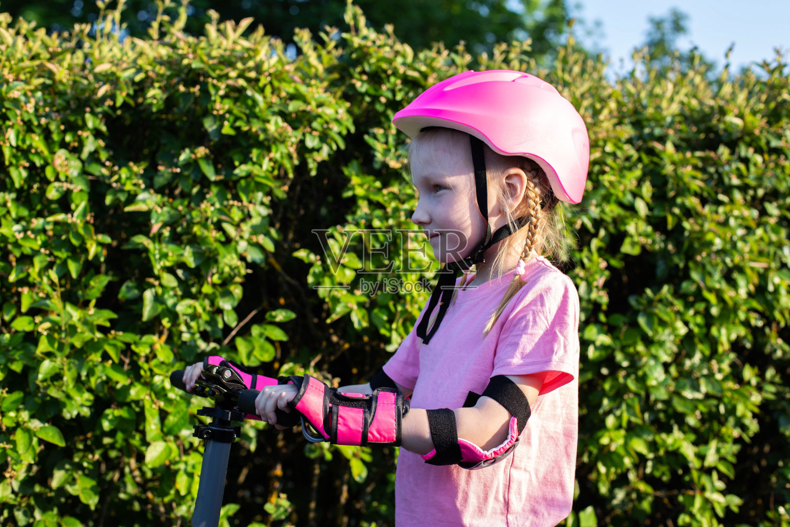 一个7岁的漂亮女孩戴着粉色的头盔,穿着防护袖,骑着摩托车站在绿色灌木丛的背景下。安全骑滑板车。照片摄影图片