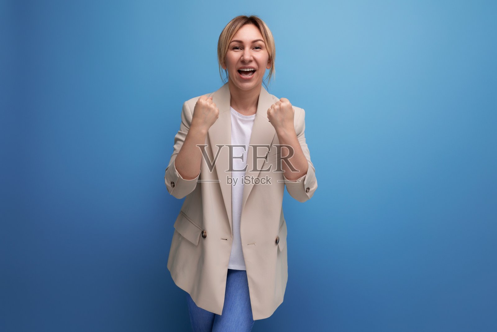 身穿米色夹克的金发女商人在蓝色背景上欢快地高呼胜利照片摄影图片