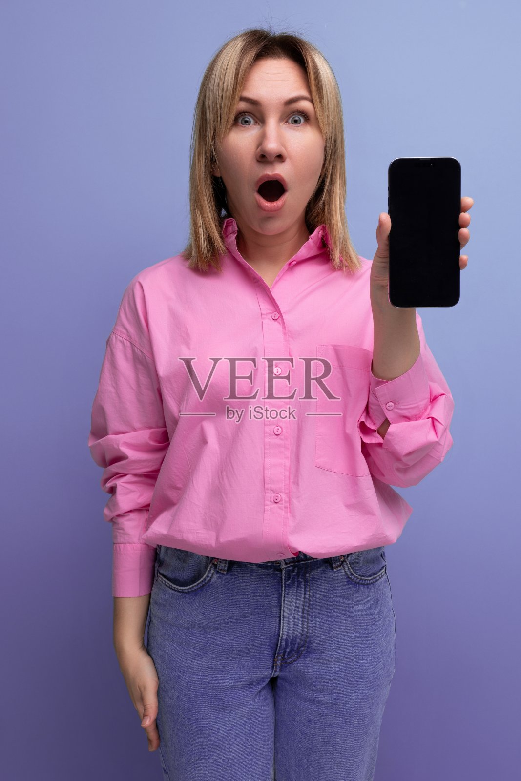 一名年轻的金发欧洲女子穿着粉色衬衫和牛仔裤,惊讶地展示着手机屏幕上的一个模型照片摄影图片