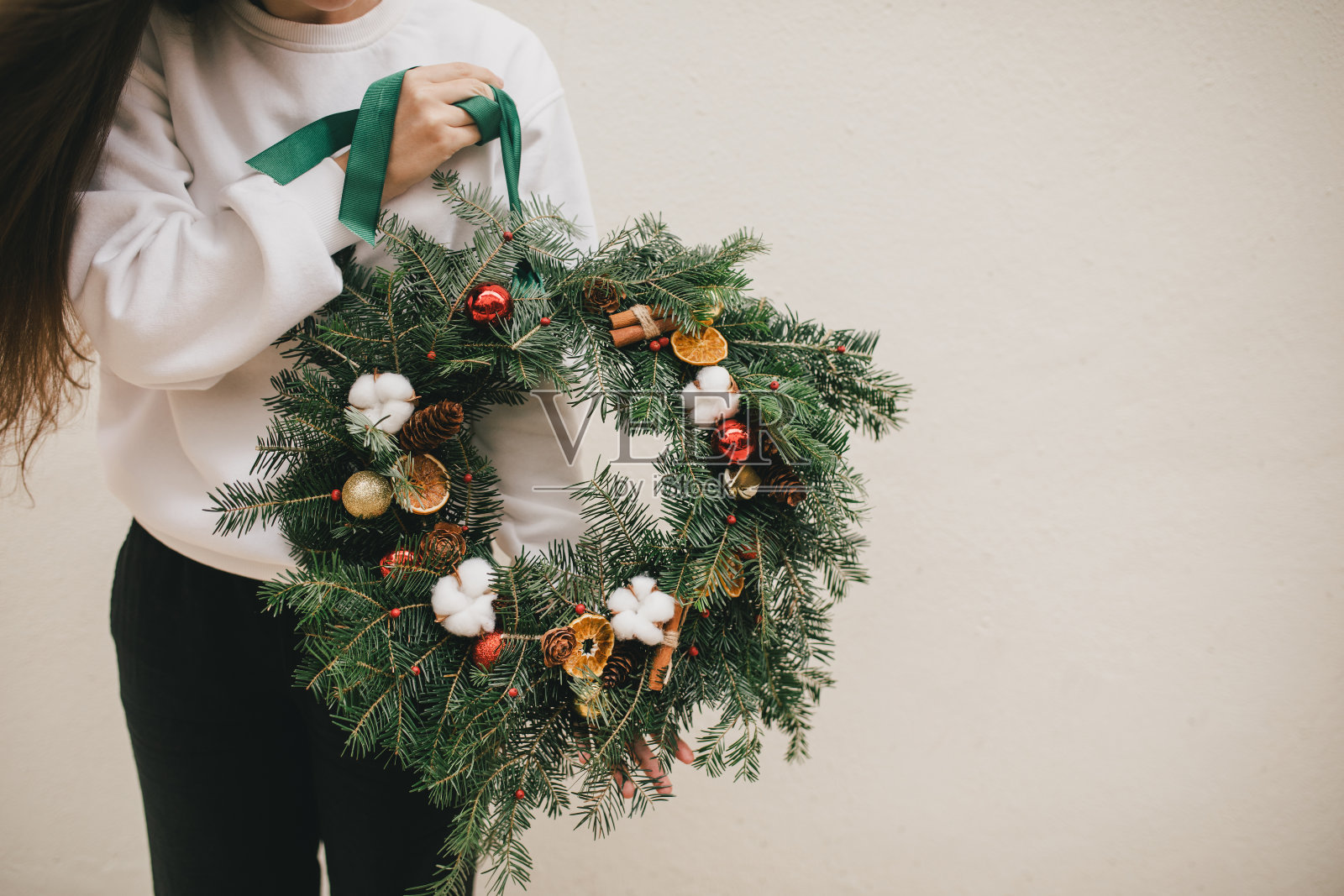 年轻女子手持手工制作的圣诞花环站在白墙旁。节日的节日装饰。照片摄影图片