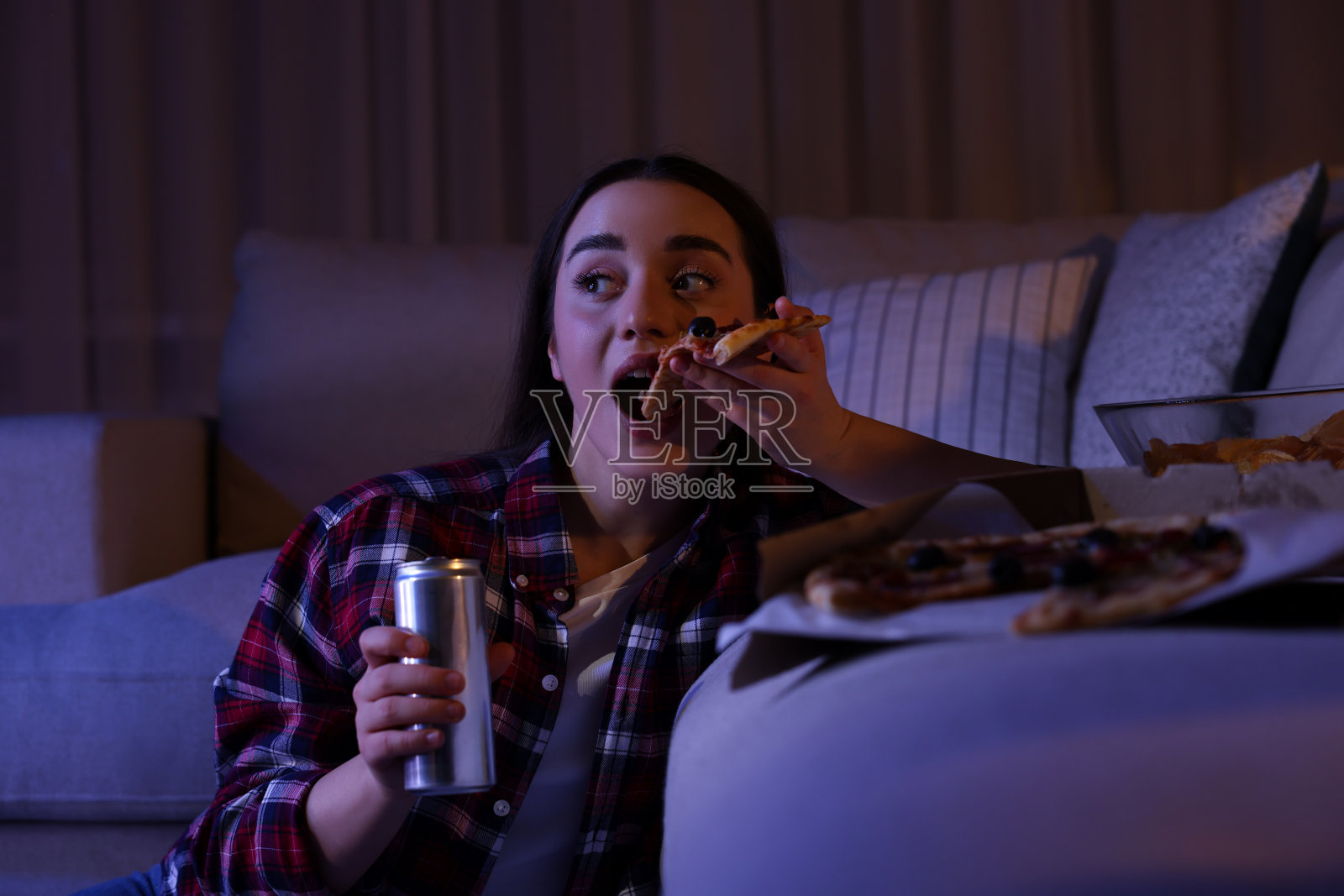 年轻女子晚上在房间里一边看电视一边吃披萨。坏习惯照片摄影图片