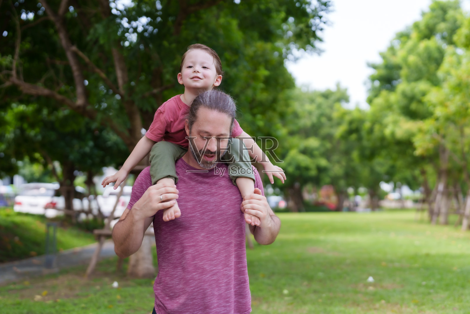 一个三岁的男孩骑在他父亲的脖子上在公园里散步。有胡子的白人男子他正在和他的儿子玩他的空闲时间。在家庭度假村草地上照片摄影图片
