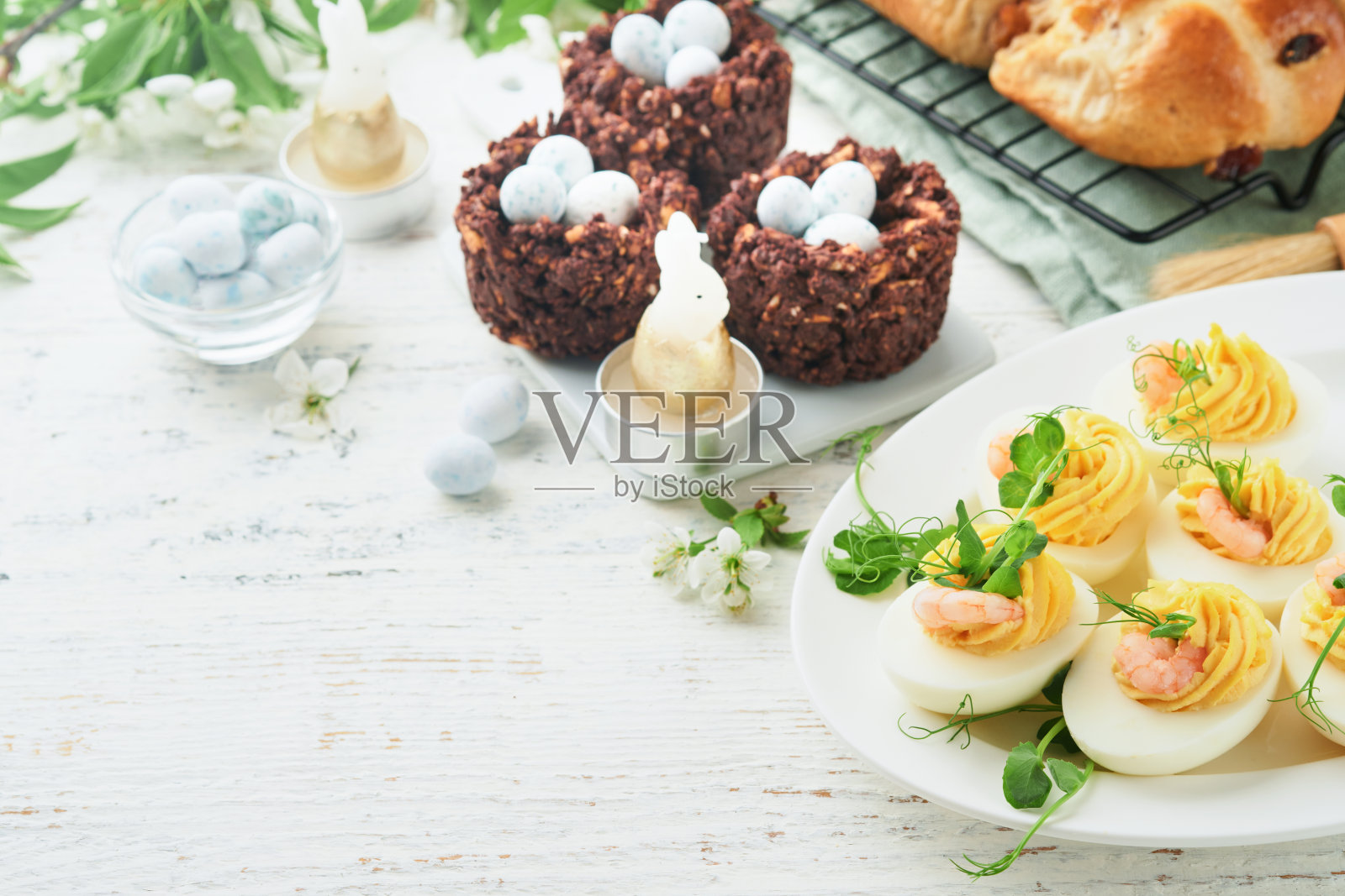 复活节餐桌上的蛋黄、虾、豌豆小菜和辣椒粉,在白色的背景上装饰新鲜的樱桃或苹果花。快乐复活节的传统菜肴概念。模拟。照片摄影图片