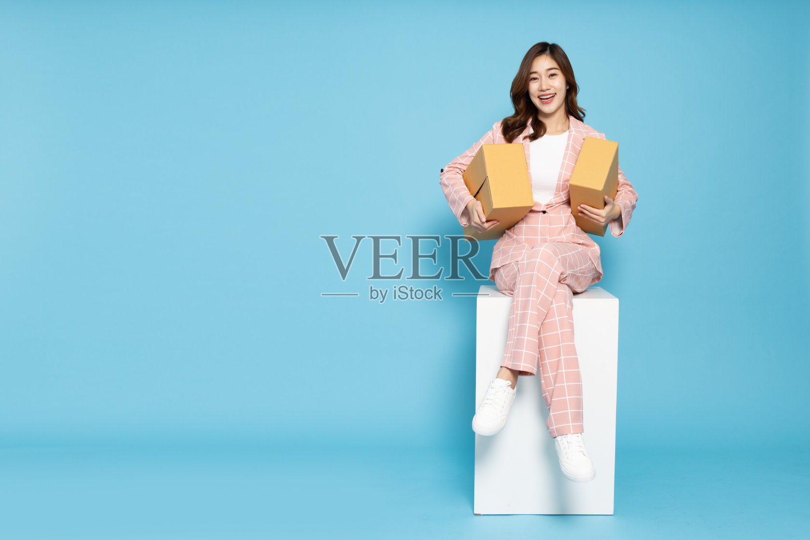 年轻的亚洲女商人拿着包裹,坐在白色的盒子上,孤立在天蓝色的背景上,快递和航运的服务理念照片摄影图片