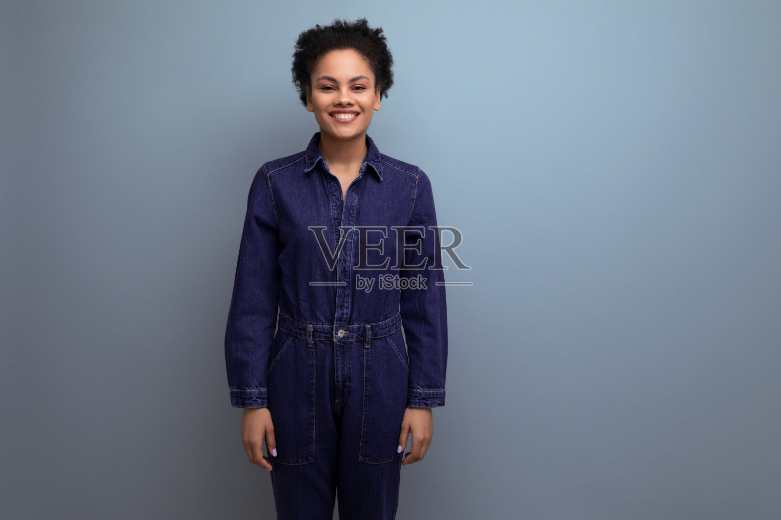 年轻积极的黑发拉丁女子,梳着非洲式头发,扎着马尾辫,穿着蓝色的时尚套装,包括连体牛仔裤和衬衫照片摄影图片