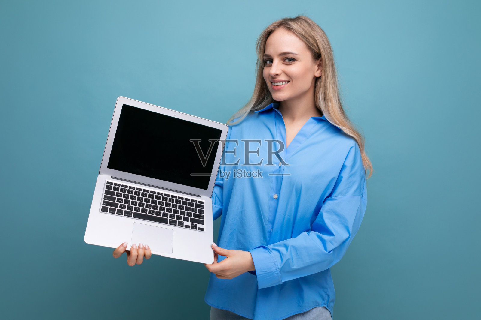 可爱的金发女孩拿着笔记本电脑,蓝色背景的空白屏幕照片摄影图片