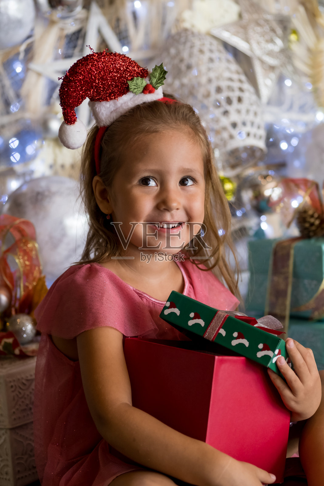 可爱的小女孩戴着圣诞老人的帽子坐在圣诞树旁边的地板上,打开礼物和礼盒。圣诞快乐,节日快乐照片摄影图片