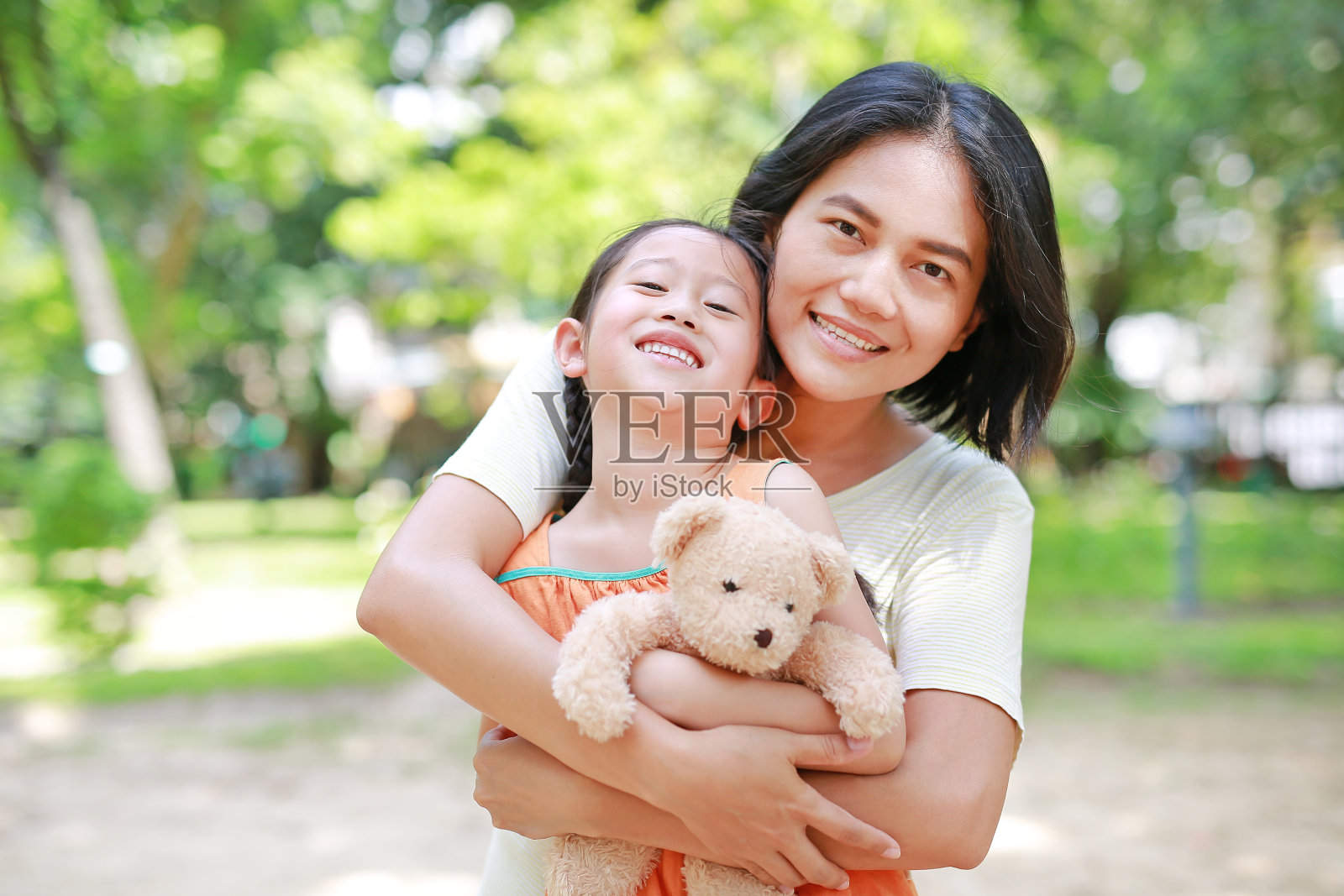 幸福的亚洲母亲在花园里抱着女儿和泰迪熊玩偶的肖像。妈妈和孩子的爱情和关系的概念。照片摄影图片