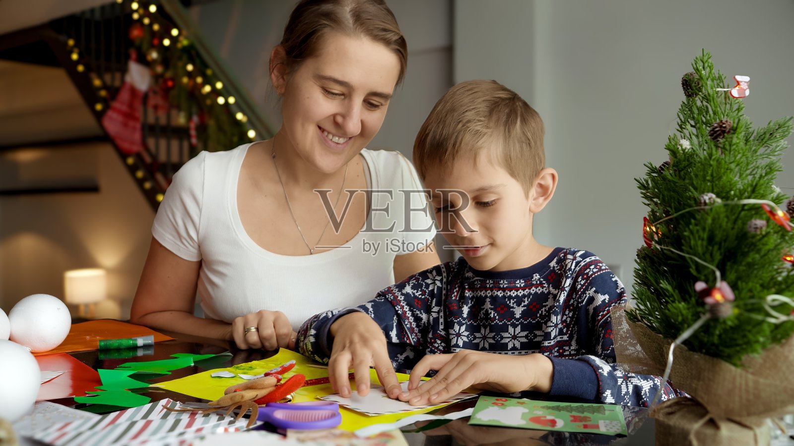 快乐微笑的母亲看着她的儿子制作传统的圣诞明信片和手工装饰照片摄影图片