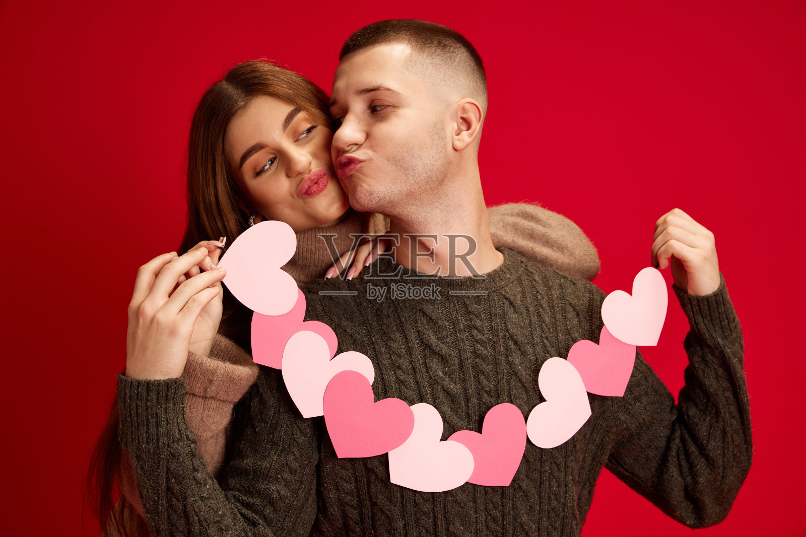 快乐,关心,爱的年轻夫妇,男孩和女孩拿着纸心花环,庆祝节日在红色的工作室背景。情人节,恋爱照片摄影图片