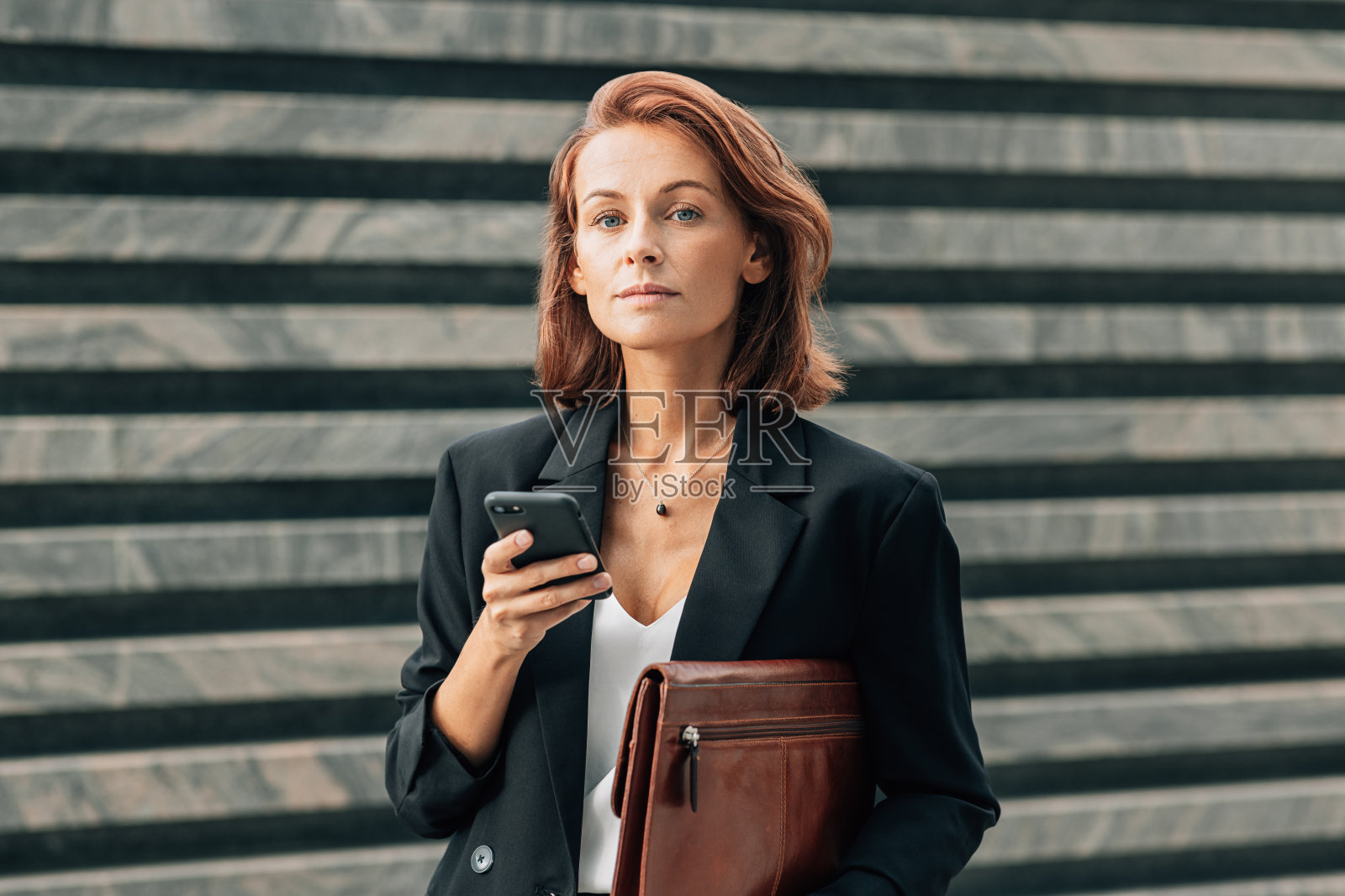 中年女商人拿着皮革文件夹和智能手机,站在户外看相机照片摄影图片