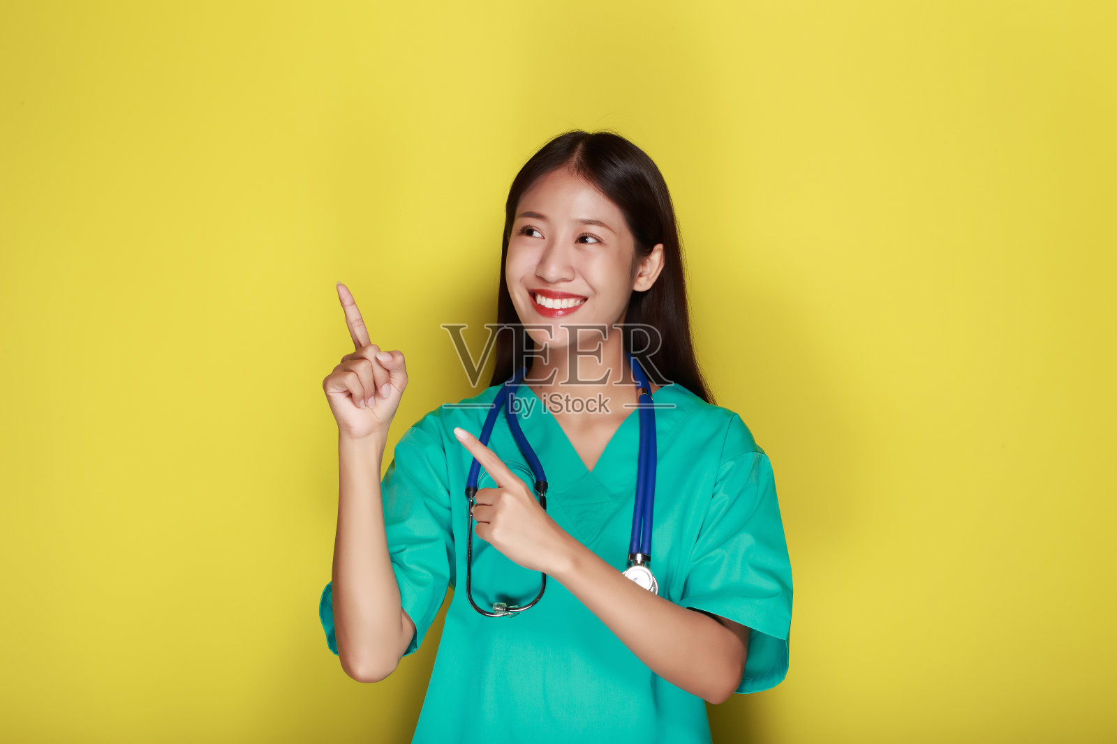 一个美丽的年轻女子在黄色背景的肖像,友好的美丽的女人穿着医生的制服,戴着听诊器指着。照片摄影图片