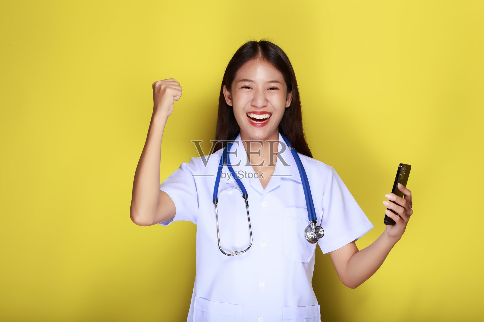 一个美丽的年轻女子在黄色背景下的肖像,亚洲妇女穿着医生的制服,戴着听诊器,拿着手机摆姿势。照片摄影图片