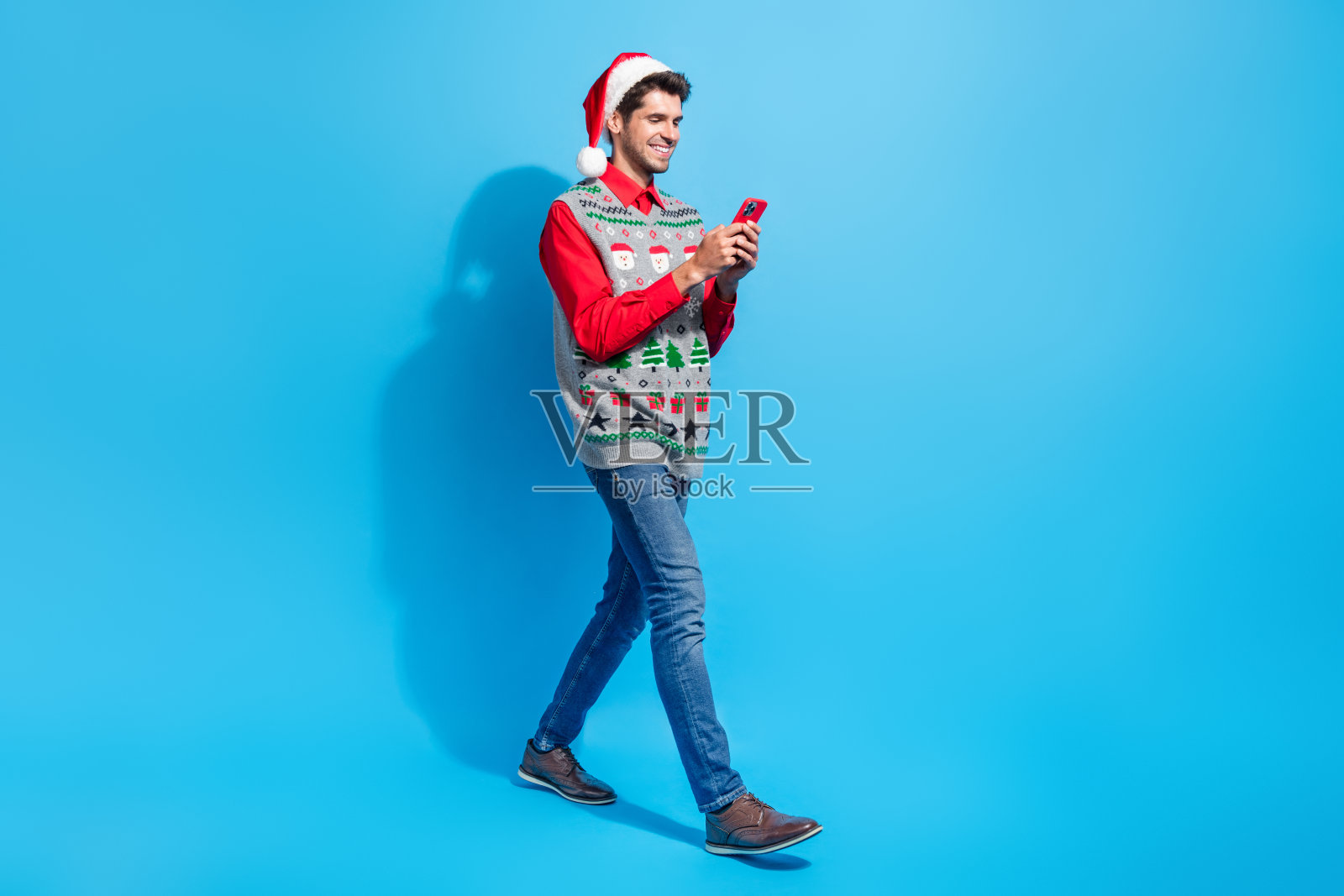 滑稽的家伙戴着圣诞帽的全身照片,看智能手机,阅读通知,在蓝色背景上孤立的空白空间照片摄影图片