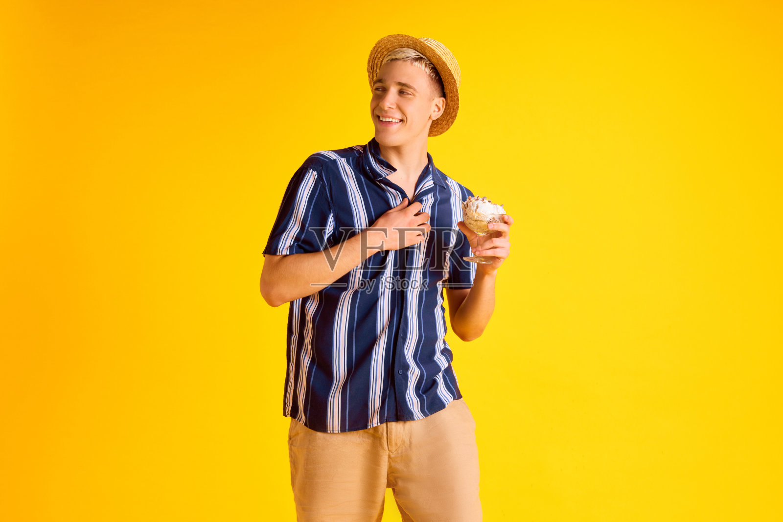 身穿条纹衬衫、短裤、头戴草帽、面带微笑的帅气年轻人,在黄色的工作室背景下吃着美味的香草冰淇淋照片摄影图片