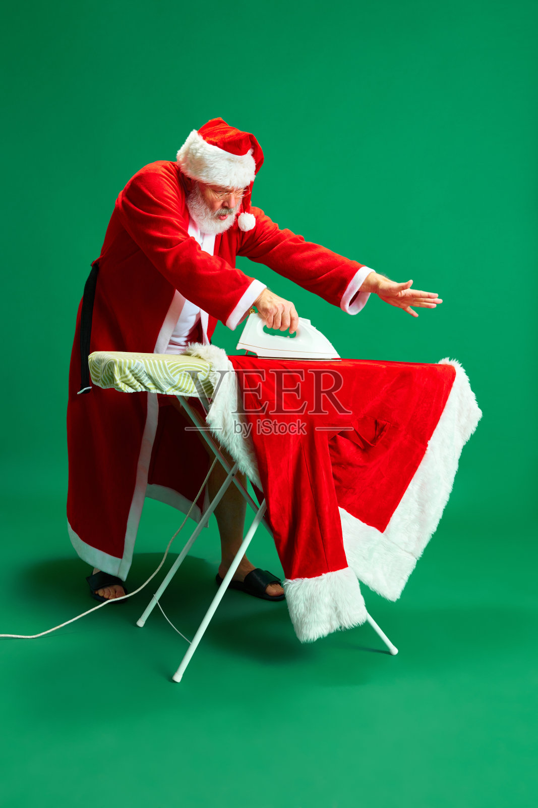 圣诞前夜,在绿色摄影棚的背景下,白发苍苍、胡须浓密的爷爷正在整理圣诞老人的节日服装。照片摄影图片