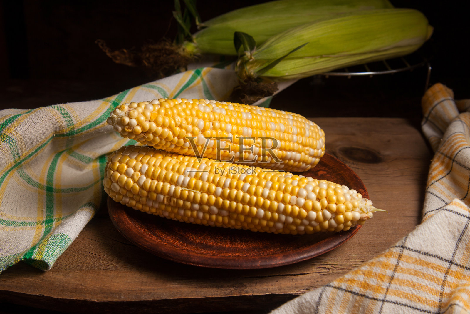 在木制背景上有两个甜玉米棒子的粘土盘子。照片摄影图片