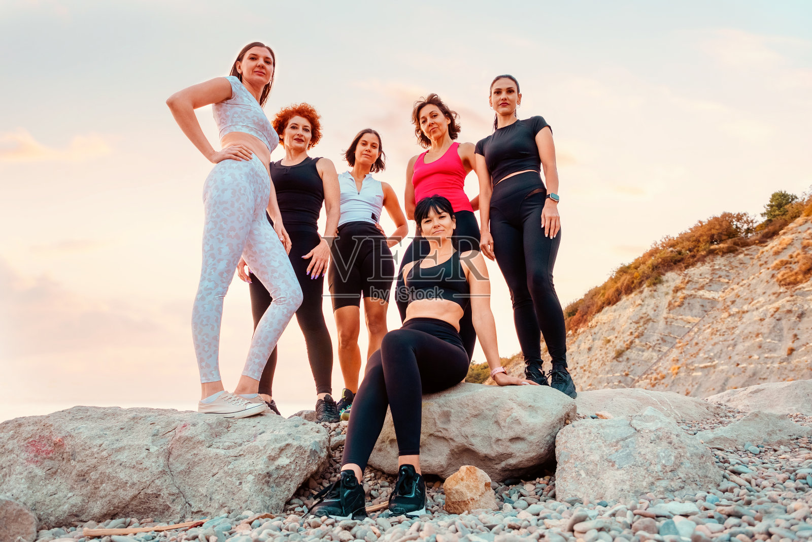 下图是一群穿着运动服的白人美女在岩石前摆姿势。背景是大海和日落。运动和女性气质的概念照片摄影图片