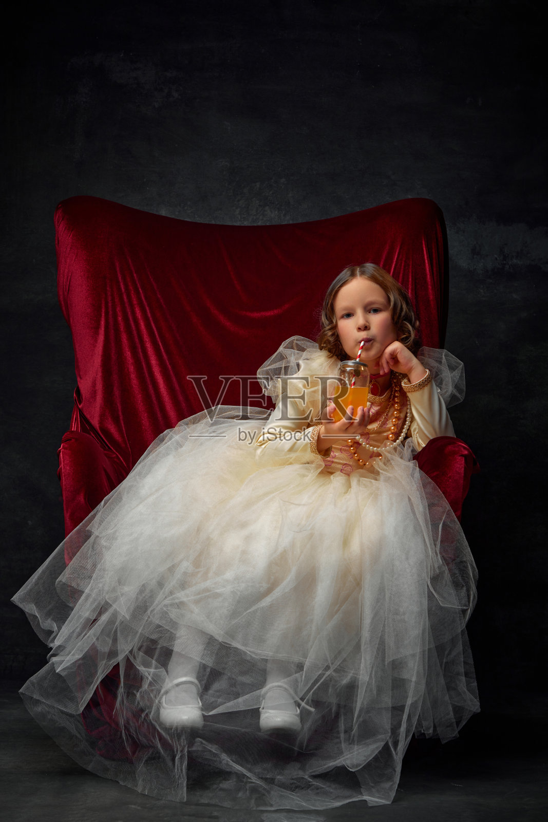 小女孩,穿着皇室成员的节日服装,坐在那里喝着苏打水,背景是黑色的复古。照片摄影图片