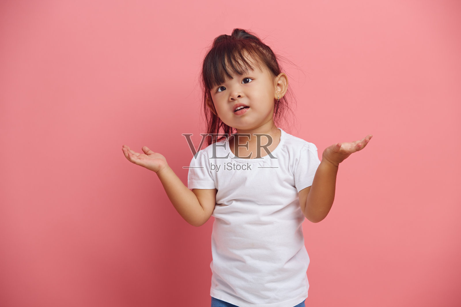 困惑的亚洲小女孩耸耸肩,露出疑问的表情,说我不知道,有一个艰难的选择,孤立地站在粉红色的背景上,穿着白色的t恤。照片摄影图片