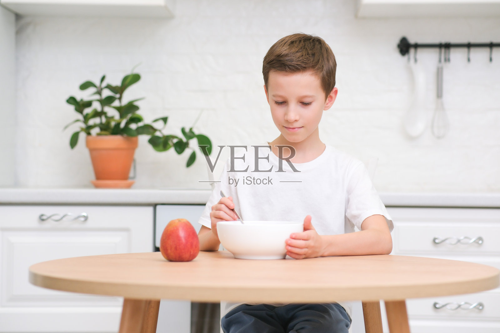 小男孩在淡白色厨房的木桌前吃着麦片配牛奶和苹果。照片摄影图片