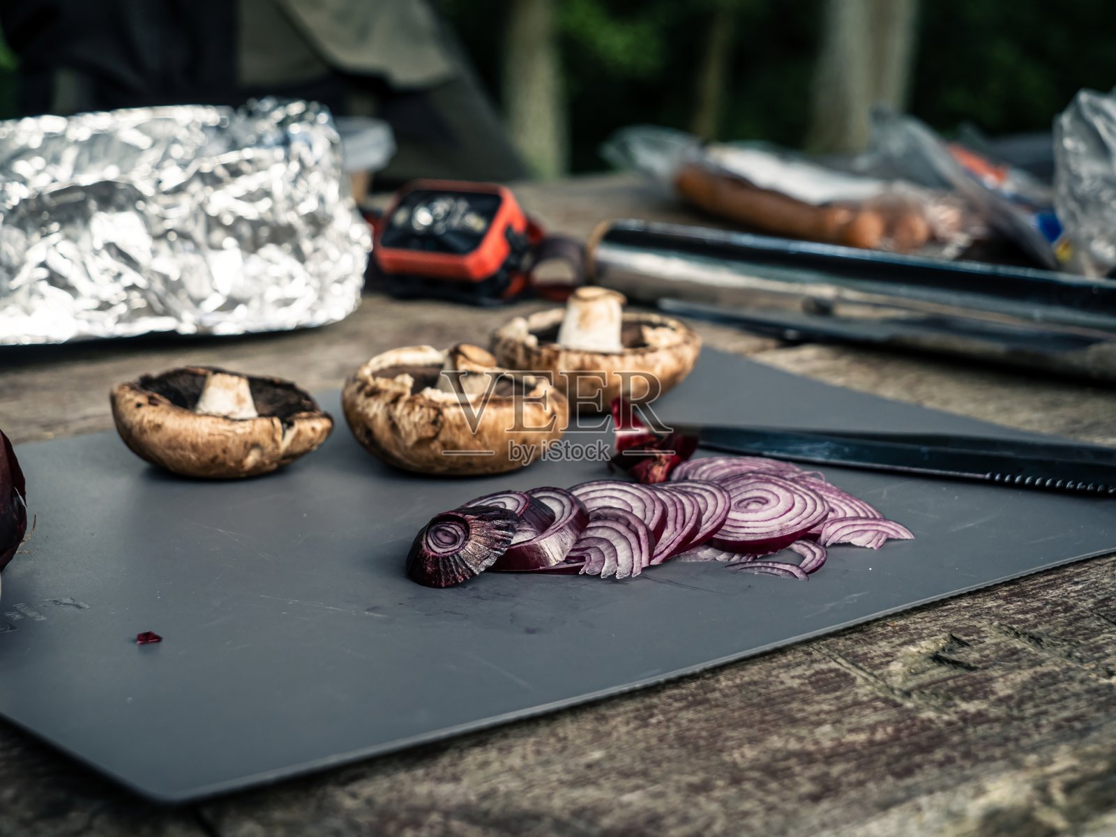 砧板上的户外木制露营桌,切红洋葱,蘑菇,大蒜,黑色猎刀照片摄影图片