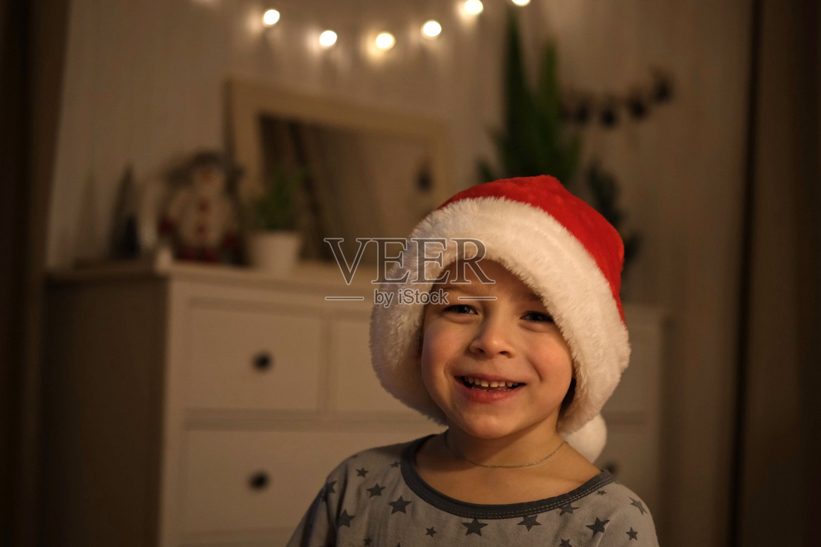 圣诞快乐。滑稽的小男孩边笑边拿着圣诞礼盒,玩得很开心。新年快乐。兴奋的小孩穿着睡衣,在床上跳来跳去。戴着红色圣诞帽跳舞的婴儿。照片摄影图片