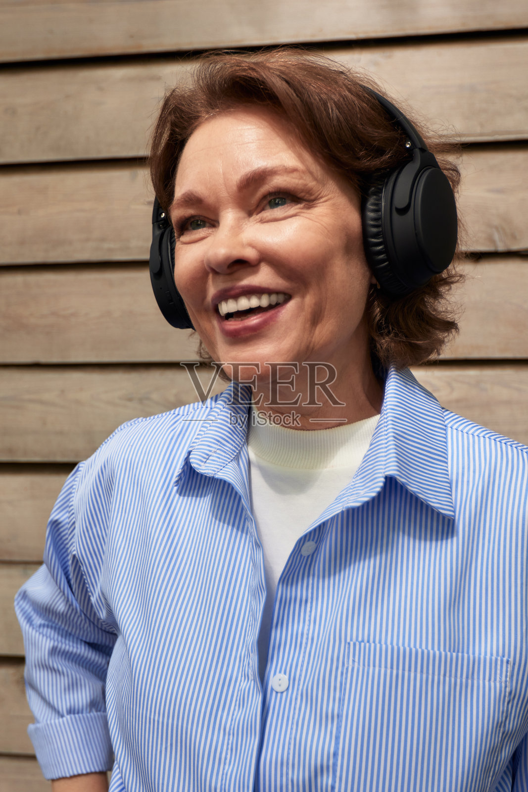 有吸引力的成熟妇女的肖像与无线耳机在墙板房子的墙壁背景。穿蓝色衬衫的黑发中年女性在听音乐。老年人积极的生活方式。照片摄影图片